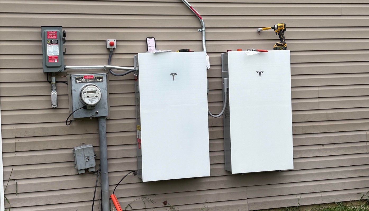 Two Tesla power wall battery units are mounted on a wall beside an electrical meter and a working tool.