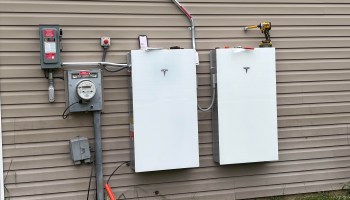 Two Tesla power wall battery units are mounted on a wall beside an electrical meter and a working tool.