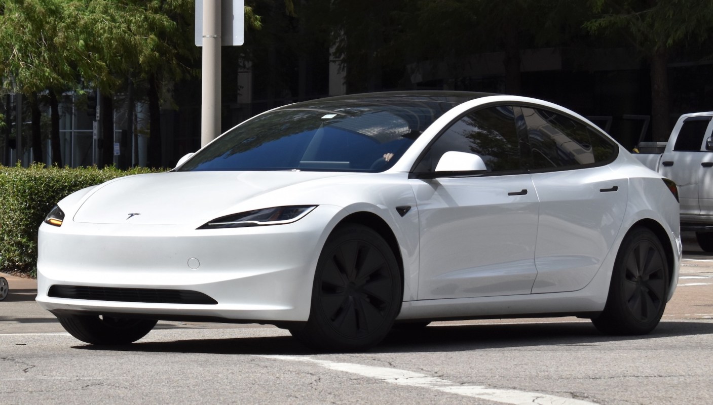 A white Tesla car parked on a street with greenery in the background.