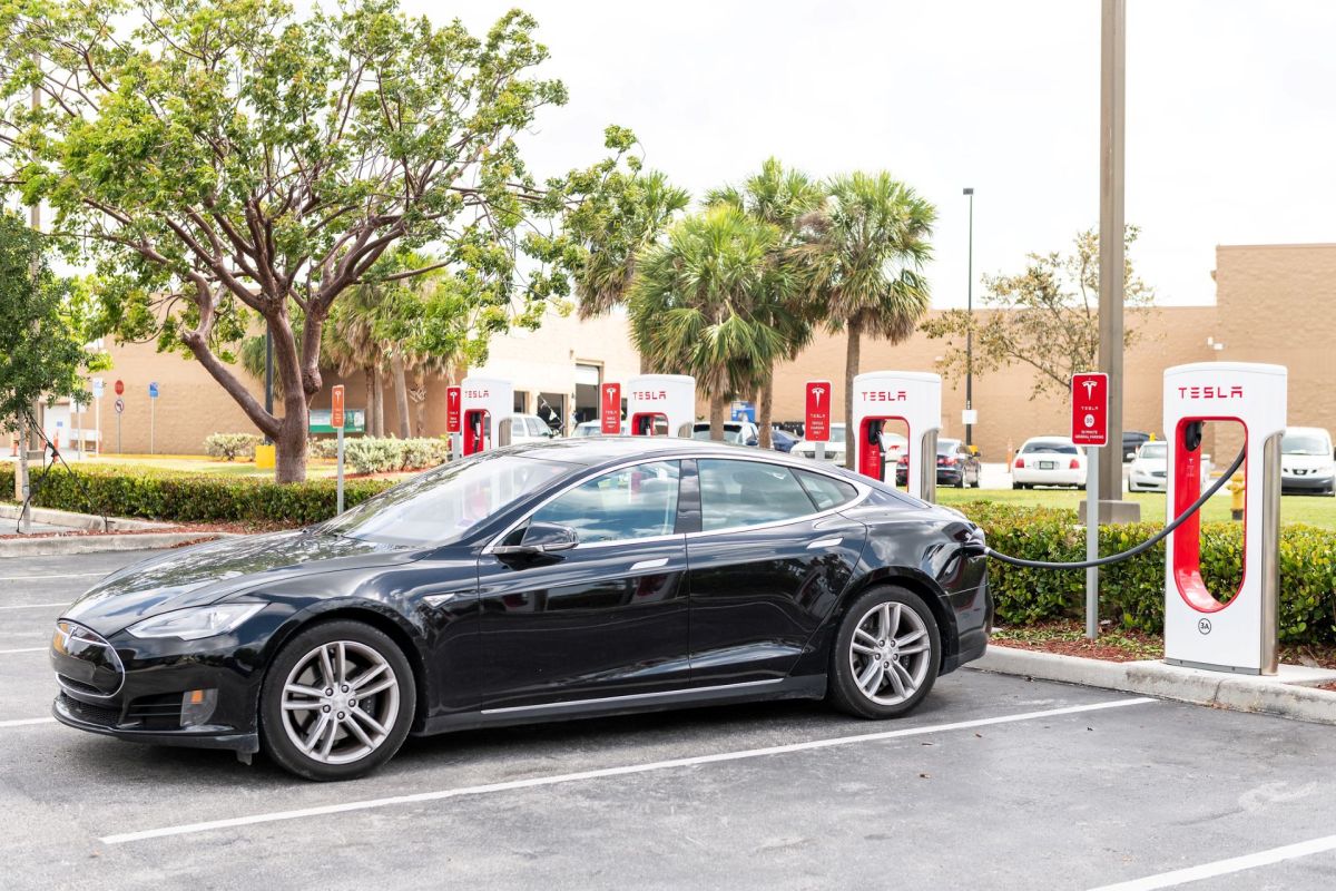 A black car is charging at a Tesla supercharger station in a parking lot.