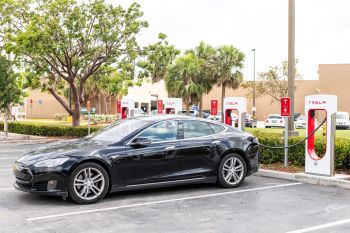 A black car is charging at a Tesla supercharger station in a parking lot.
