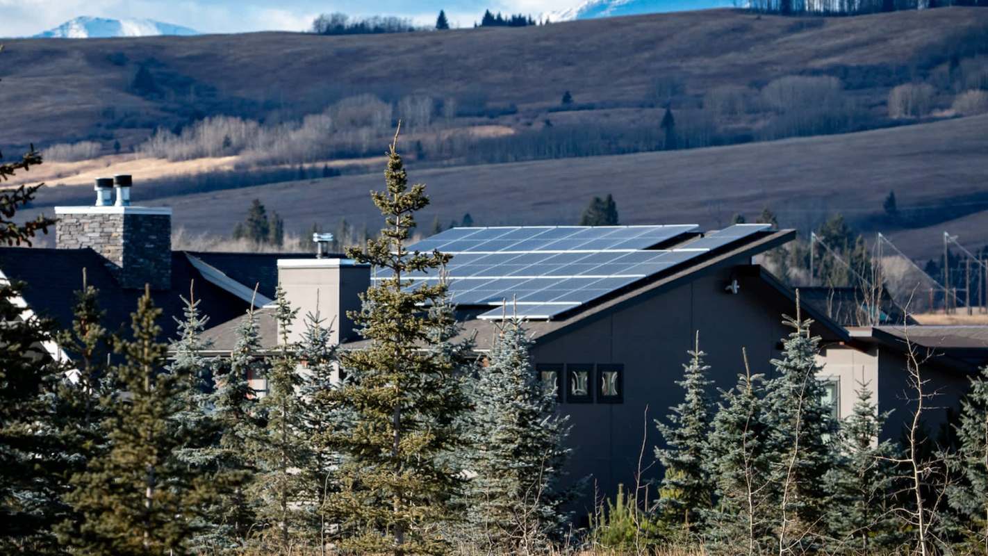 A house with solar panels on the roof surrounded by evergreen trees and rolling hills in the background.