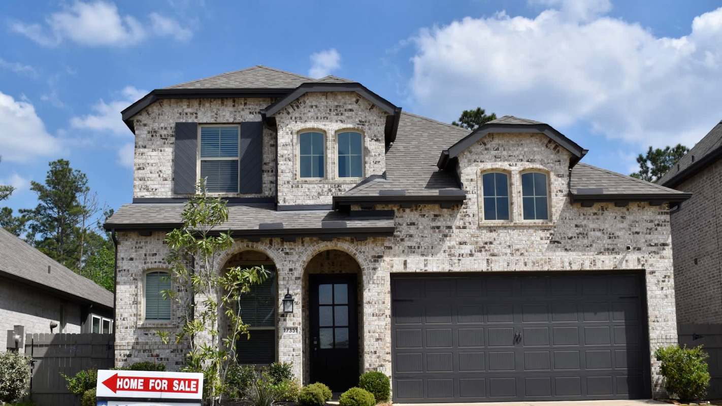 A two-story brick house with a garage and a "Home For Sale" sign in the front yard.