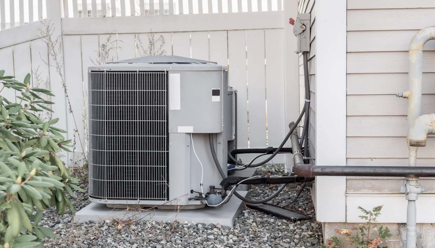 An outdoor air conditioning unit placed next to a house, surrounded by gravel and plants.