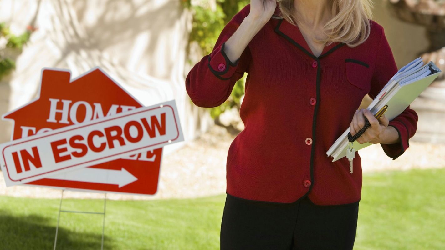 A woman holds files and a phone near a "Home for Sale" sign with "In Escrow" on a lawn.