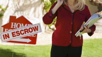A woman holds files and a phone near a "Home for Sale" sign with "In Escrow" on a lawn.