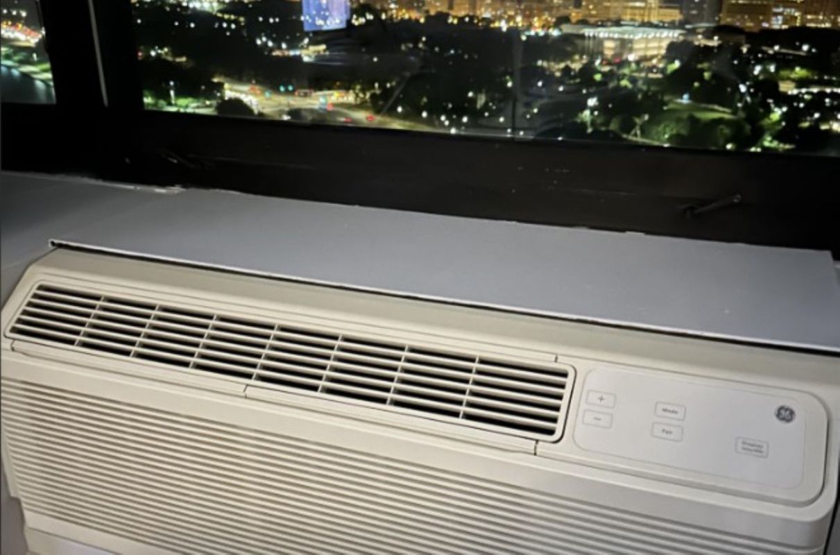 An air conditioning unit placed near a window overlooking a city skyline at night.