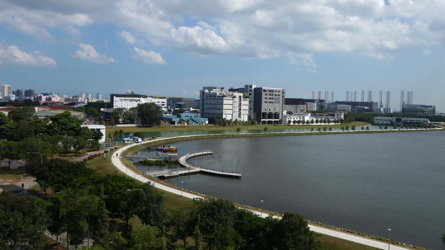 A waterfront view featuring a pathway, green spaces, buildings, and a blue sky with clouds.
