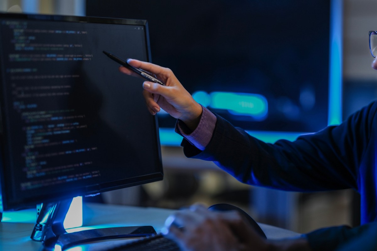 A person points at code on a computer screen while typing on a keyboard.