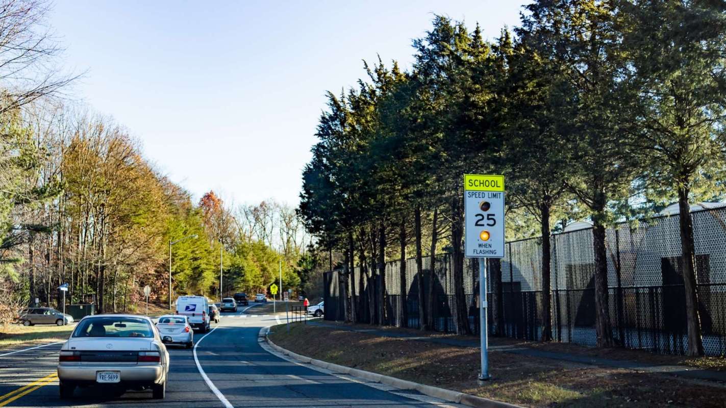 A road with vehicles and a school zone speed limit sign indicating 25 mph when flashing, bordered by trees.