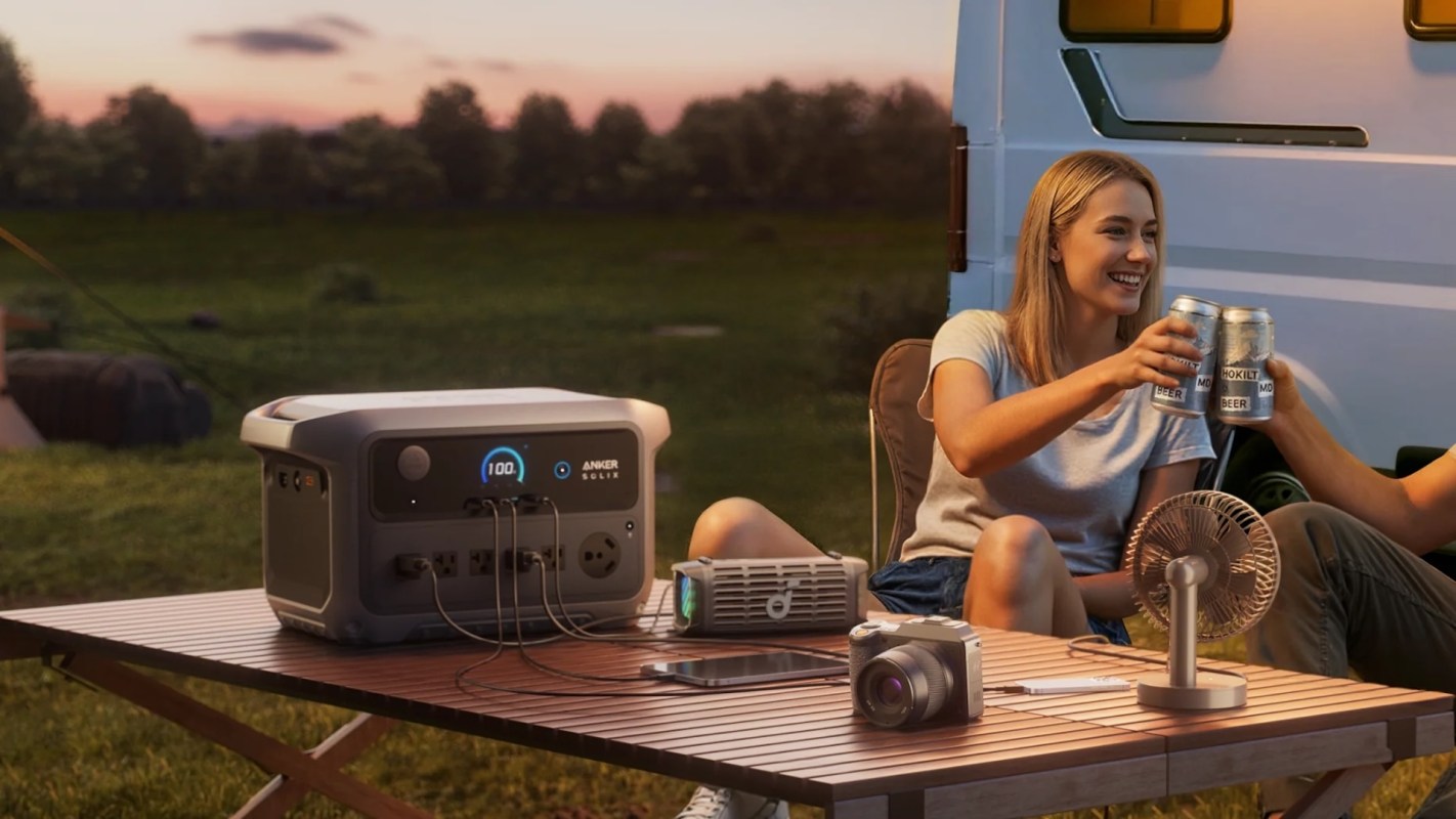 Two people toast with cans of beer while sitting at a table with a portable power station and a fan in the background.