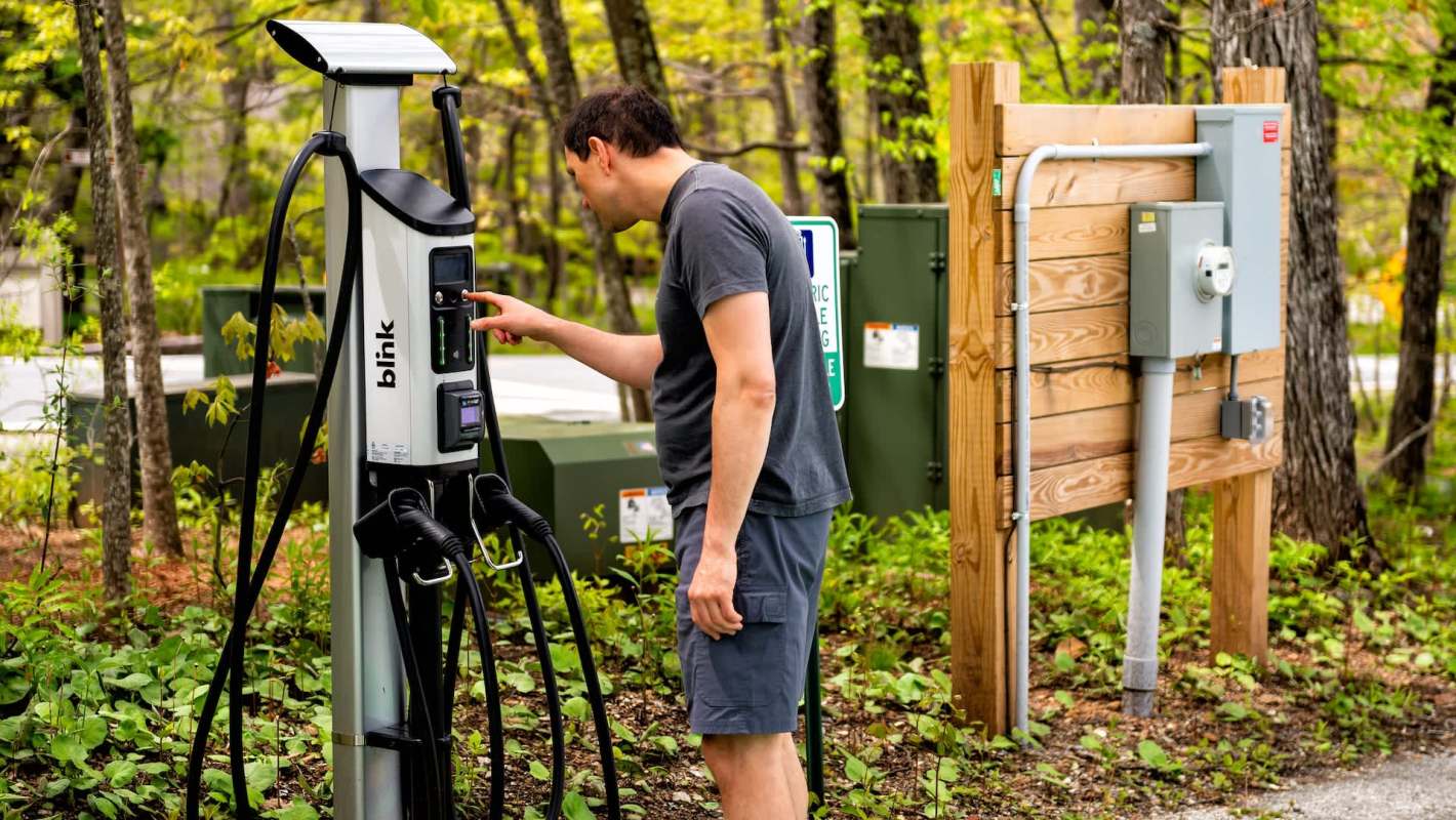 A man interacts with an electric vehicle charging station surrounded by greenery.