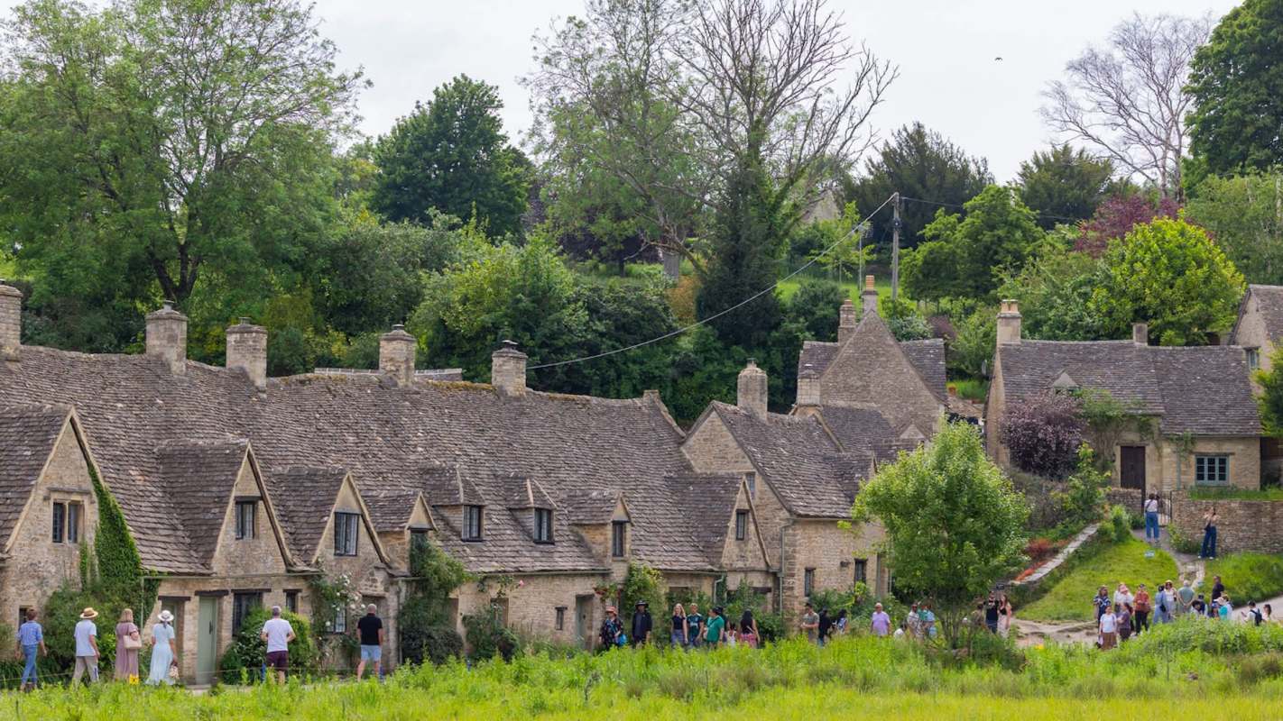A line of cottages in Arlington Row in Bibury, England, with visitors walking along a path in a green landscape.