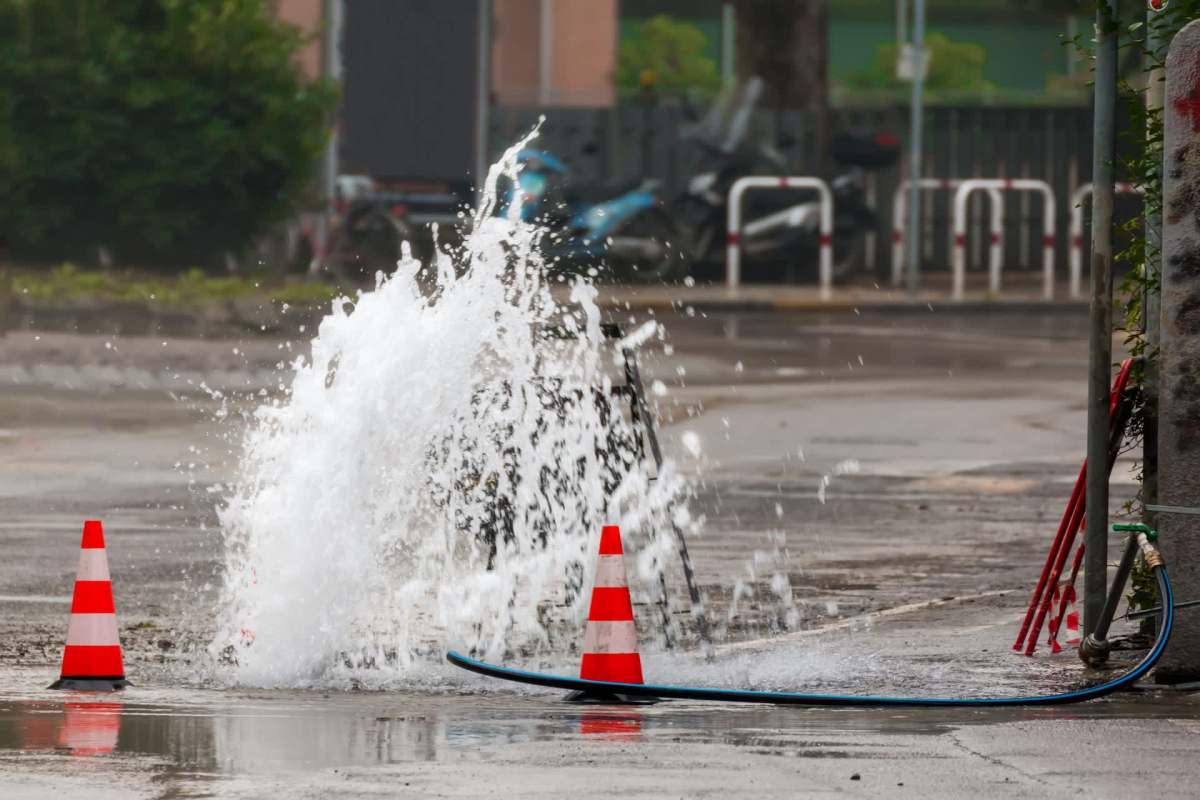 Water erupts from the ground near traffic cones on a wet street.
