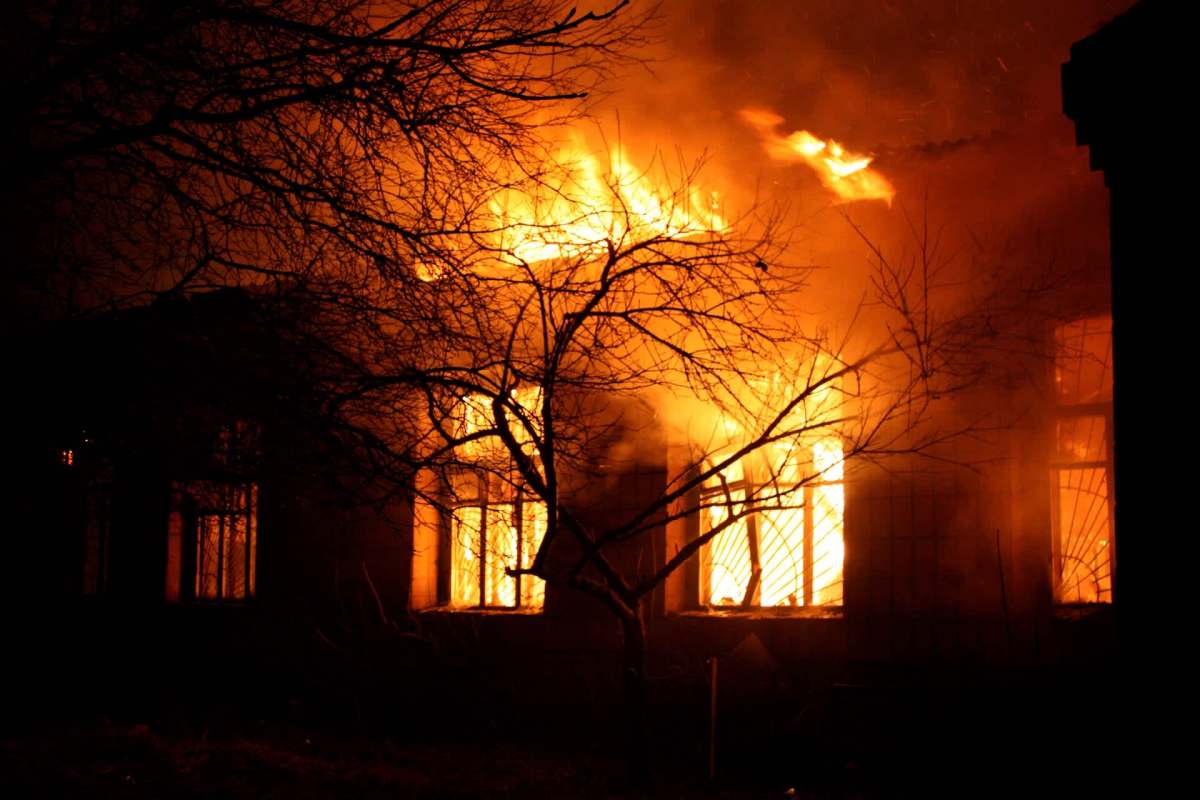 A building engulfed in flames with dark smoke rising, framed by bare tree branches.