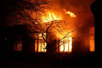 A building engulfed in flames with dark smoke rising, framed by bare tree branches.