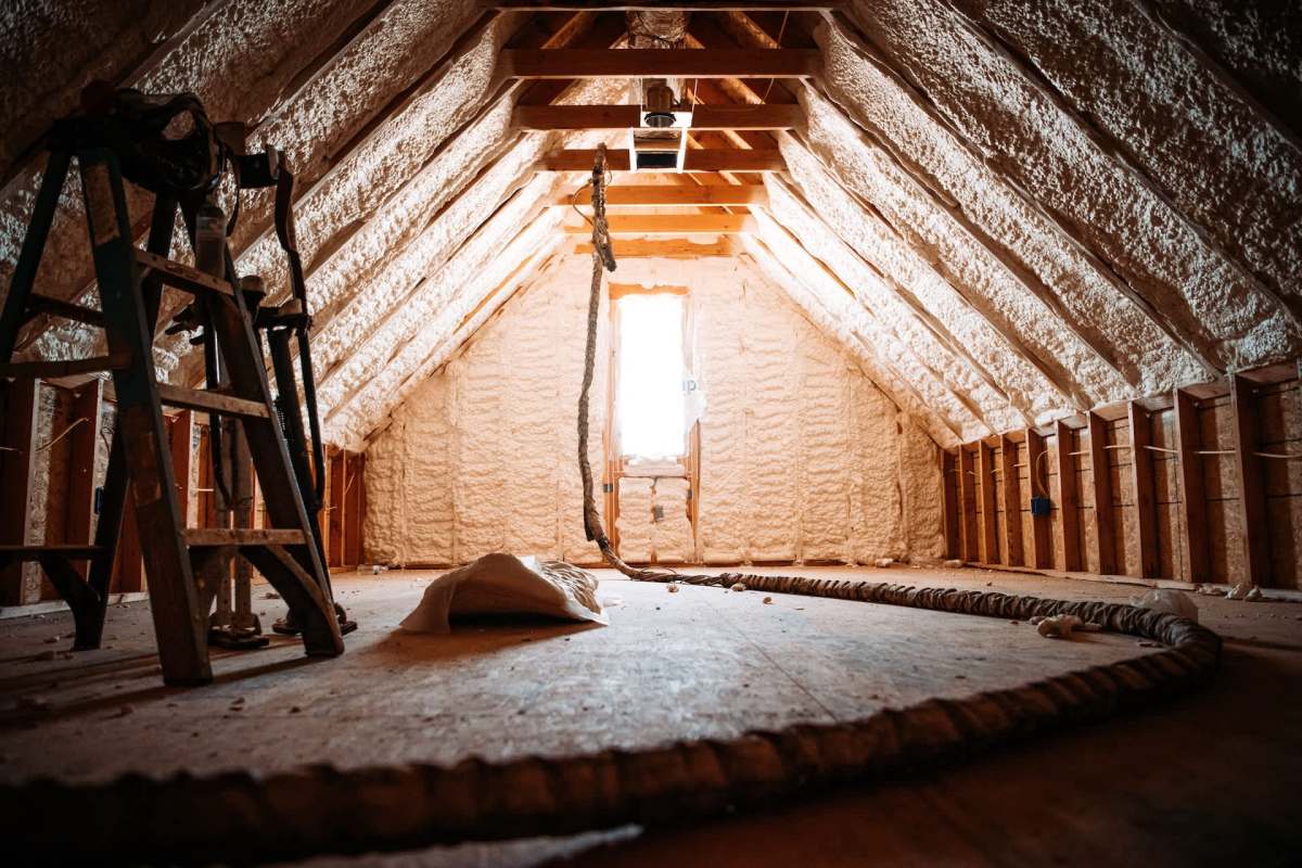 An empty attic with insulation on the walls, a ladder, and a window allowing natural light.