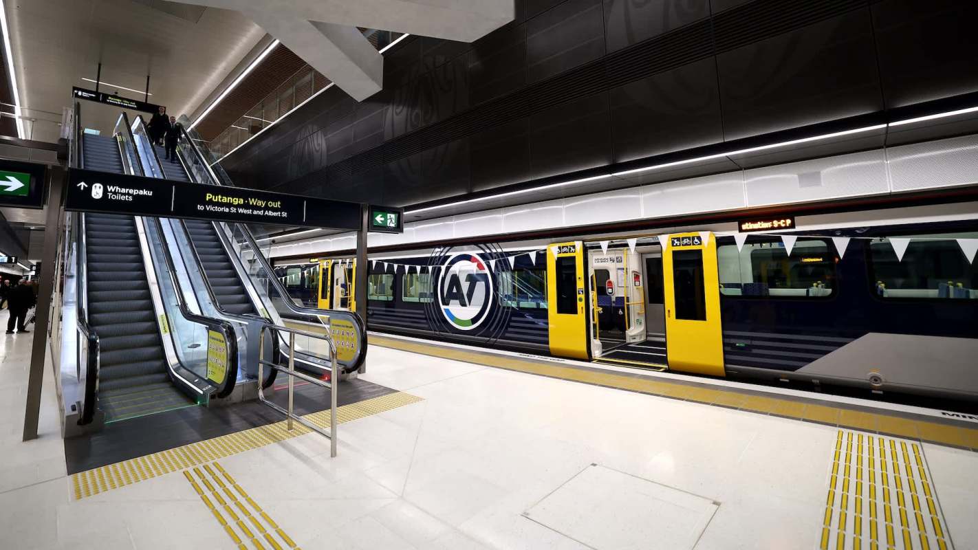 An escalator leads to a train platform featuring a yellow and blue train with digital signage.