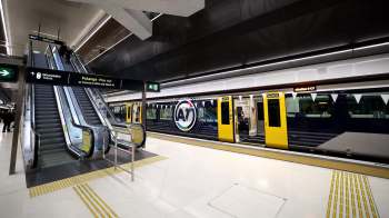 An escalator leads to a train platform featuring a yellow and blue train with digital signage.