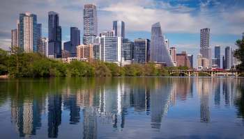An Austin, Texas, skyline of modern skyscrapers reflected in a calm body of water with greenery in the foreground.