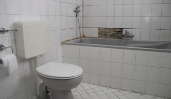 A bathroom with a toilet, a bathtub, and a wall showing damage and mildew.