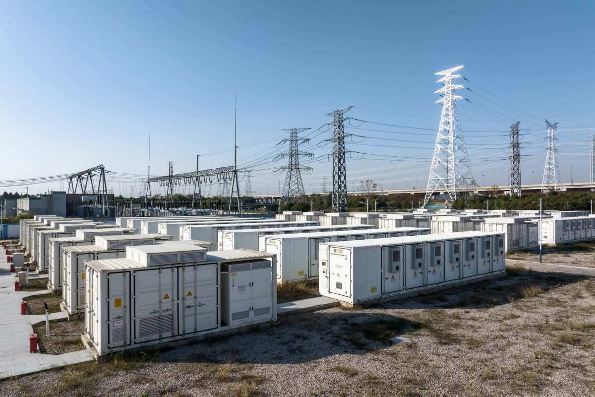 Battery storage units are lined up in an open area, with power lines and a substation in the background.