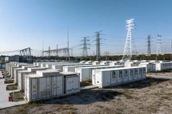 Battery storage units are lined up in an open area, with power lines and a substation in the background.