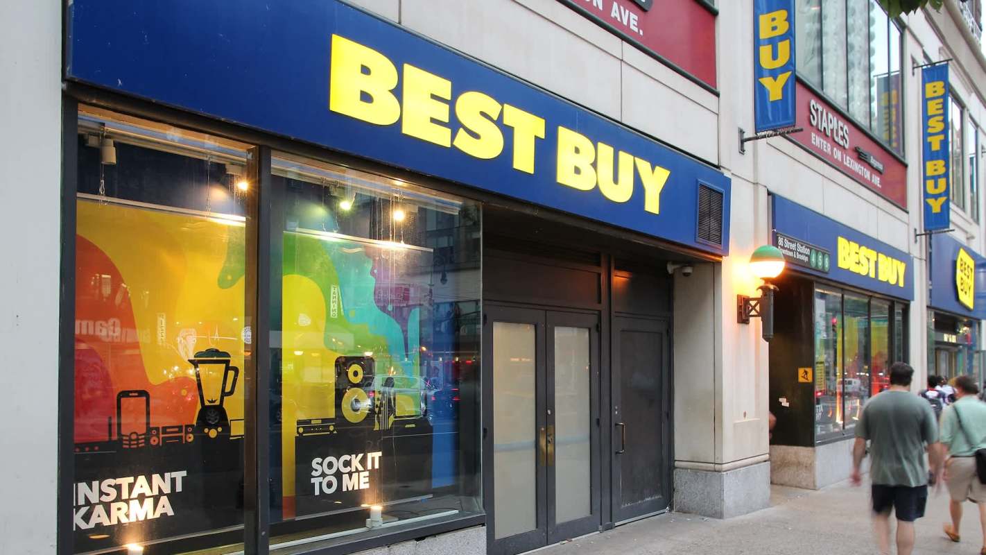 A Best Buy store with colorful window displays and pedestrians walking by.