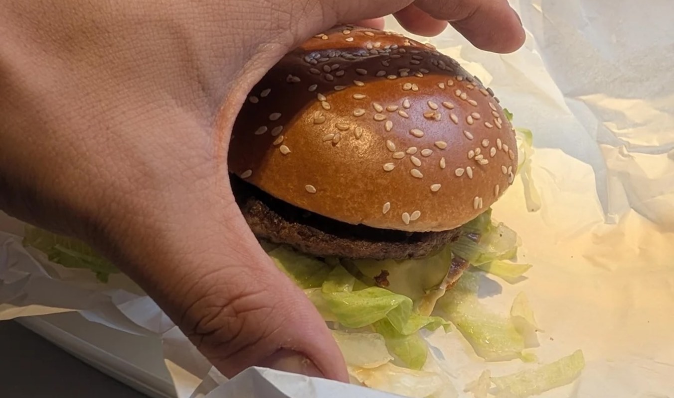 A hand holds a sesame seed bun with lettuce peeking out, resting on white paper.