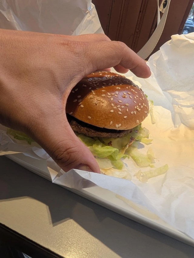 A hand holds a sesame seed bun with lettuce peeking out, resting on white paper.