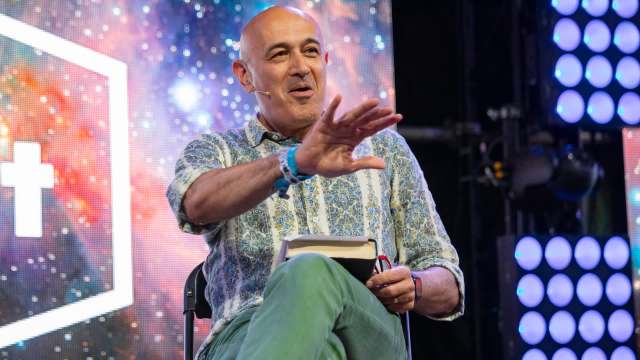 Physicist Jim Al-Khalili sits on a chair, gesturing while holding a book, with a colorful, starry background.