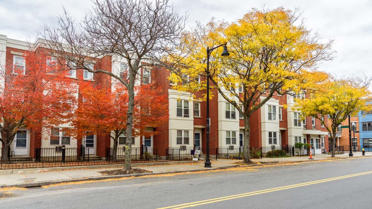 A row of buildings beside a street lined with trees displaying autumn foliage in red and yellow.