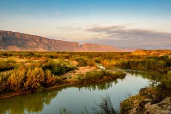 A river flows through a landscape with mountains and vegetation under a clear sky.