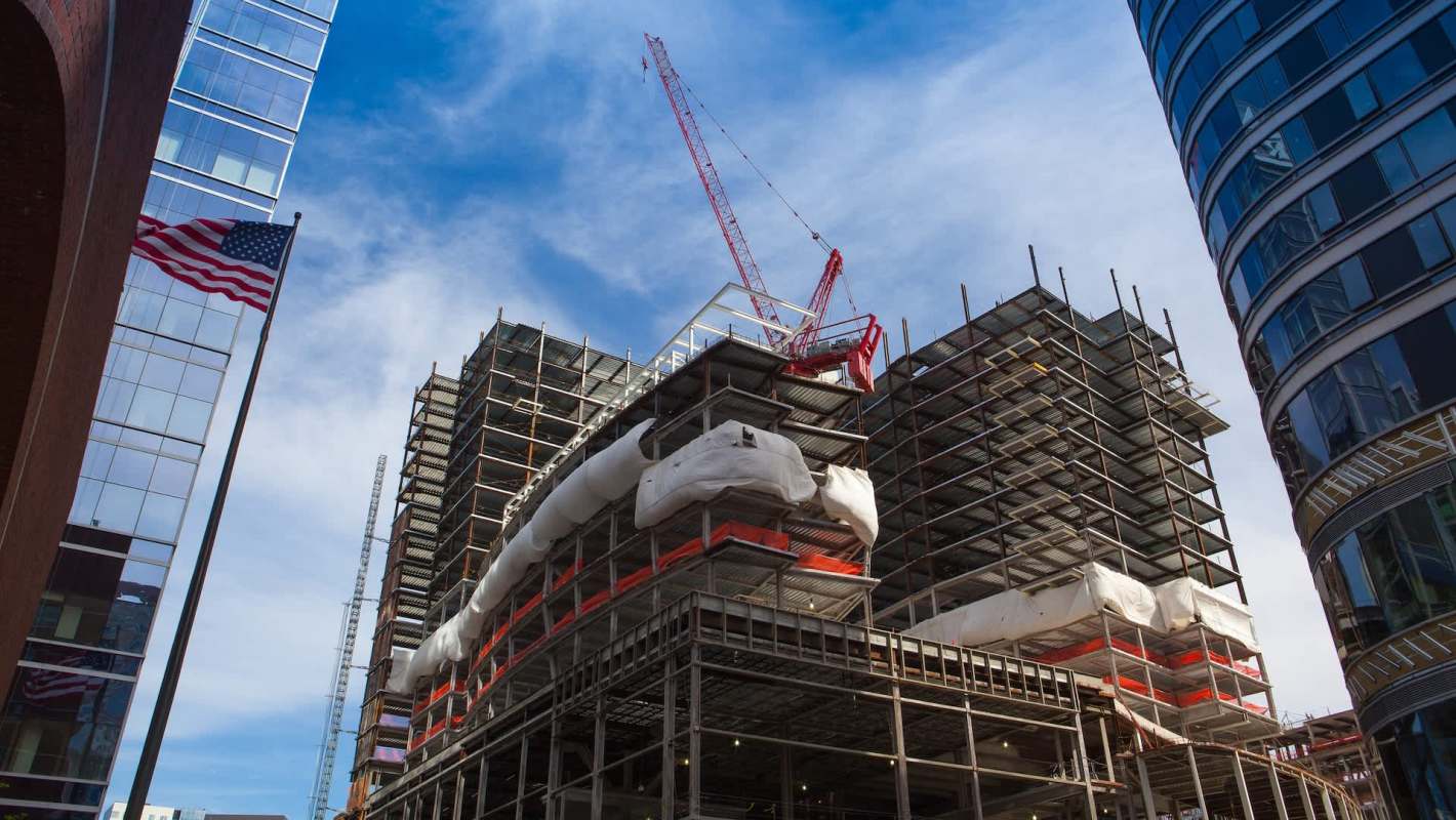 A construction site with cranes and scaffolding between modern buildings under a blue sky.