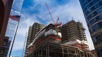 A construction site with cranes and scaffolding between modern buildings under a blue sky.