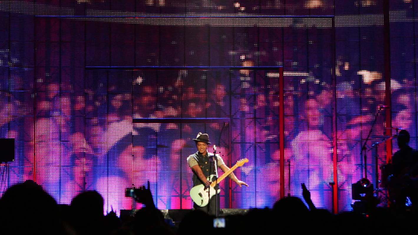 Bruno Mars on stage during his concert, with fans visible on a video screen behind.
