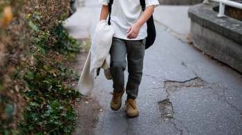 A person walks along a path, which includes cracks, holding a light-colored jacket with greenery on the side.