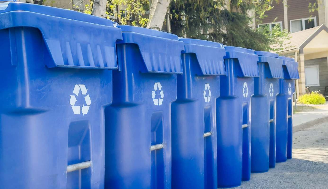Multiple blue recycling bins lined up on a street with a recycling symbol on each.