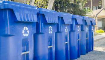 Multiple blue recycling bins lined up on a street with a recycling symbol on each.