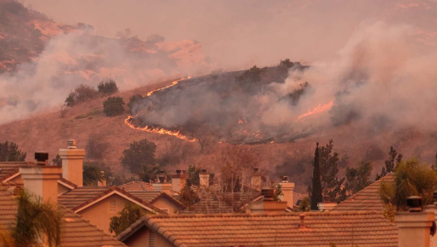 A hillside is engulfed in flames and smoke, with rooftops visible in the foreground.