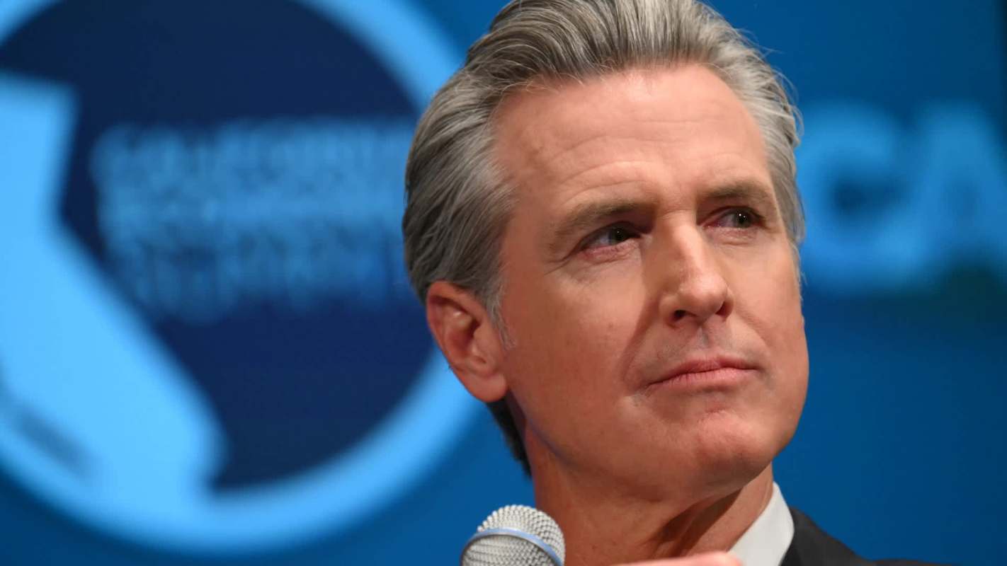 California governor Gavin Newsom speaking into a microphone with a blue background and a logo visible.
