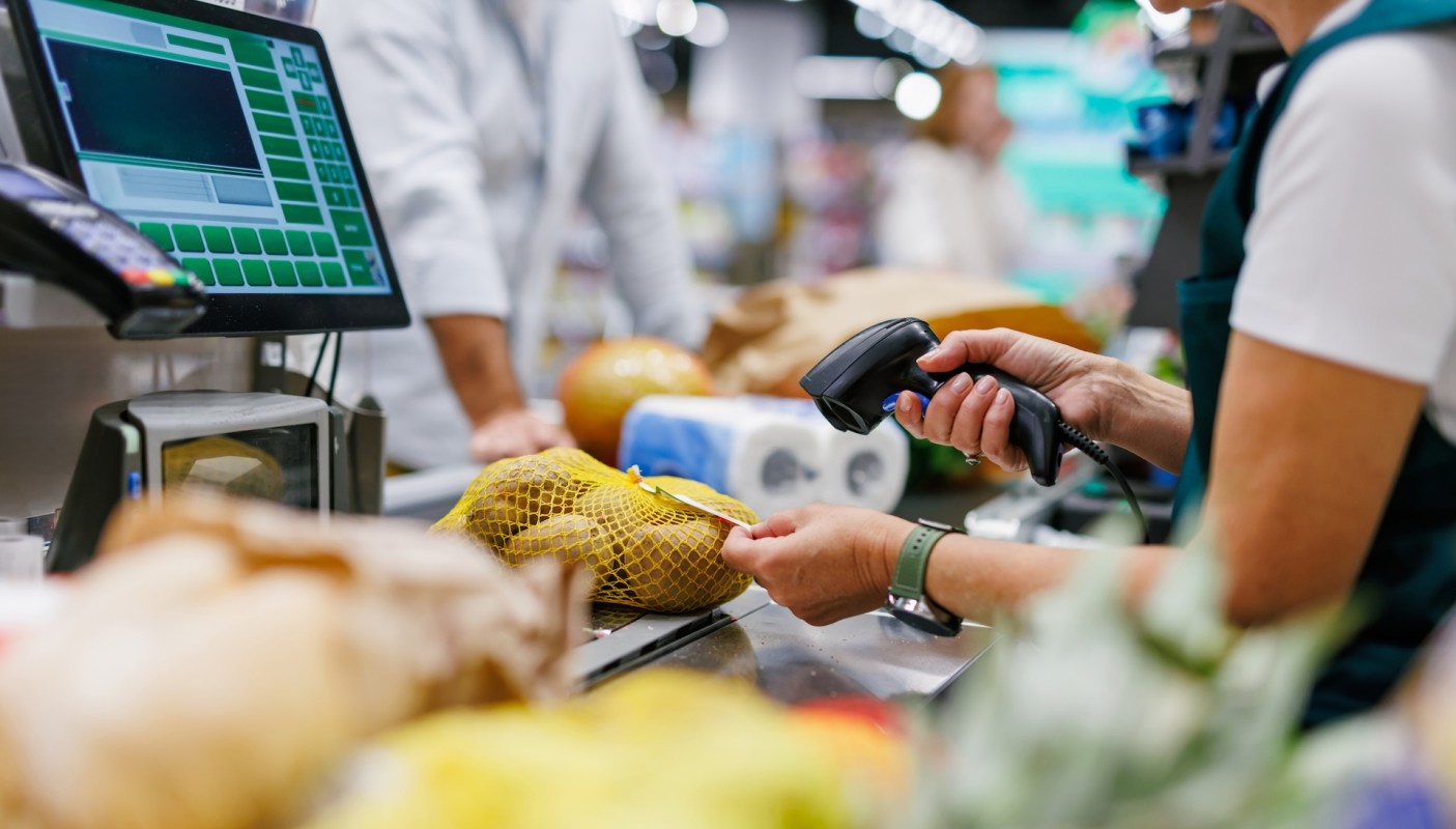 A cashier scans items at a checkout counter with a digital register and bags of groceries nearby.