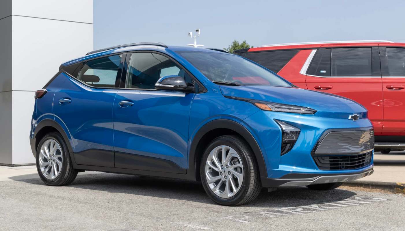 A blue Chevrolet Bolt parked in a car lot with a red vehicle in the background.