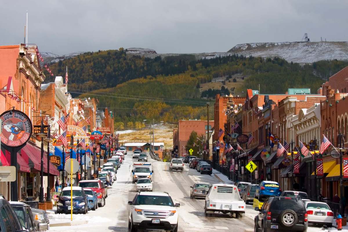 A street lined with shops and flags, with vehicles parked along the snowy road and mountains in the background.