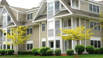 A multi-story building with beige siding, white trim, and small yellow trees in the foreground.