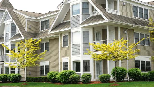 A multi-story building with beige siding, white trim, and small yellow trees in the foreground.