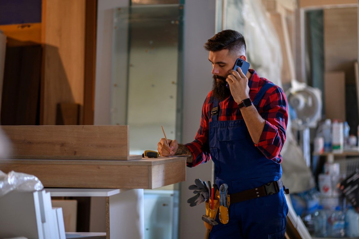A man in work attire holds a phone while writing on a notepad at a construction site.