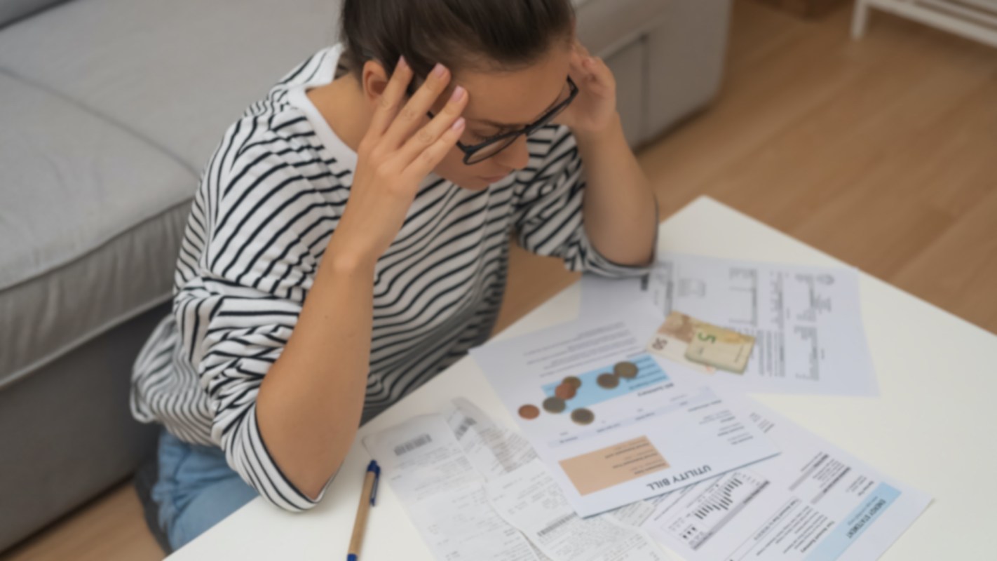 A woman sits at a table surrounded by bills, coins, and receipts, appearing stressed.