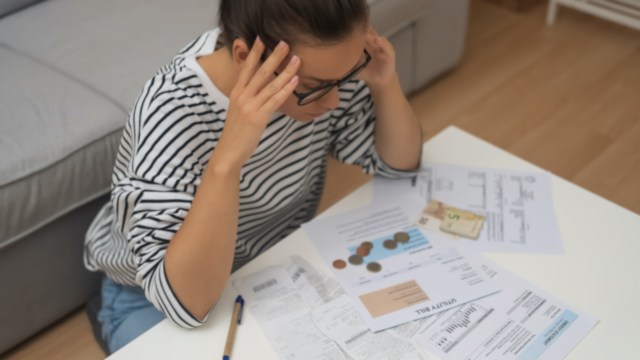 A woman sits at a table surrounded by bills, coins, and receipts, appearing stressed.