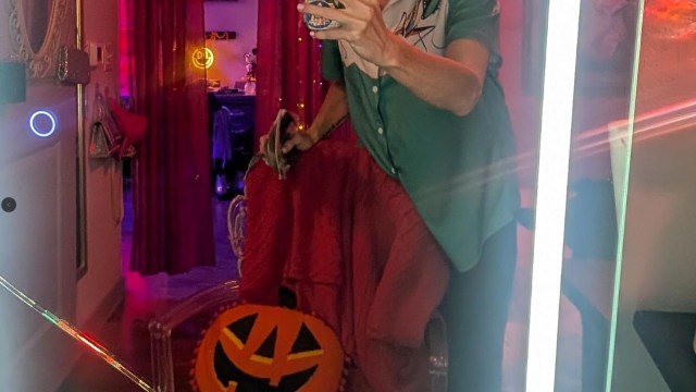 A person holds a phone in front of a cracked mirror, with a pumpkin decoration and colorful lights in the background.
